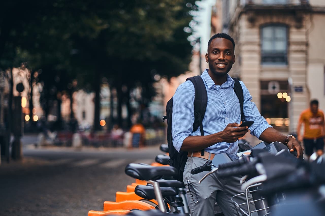 Homem negro sorridente, vestindo calça e blusa social com uma mochila nas costas. Ele aluga uma bicicleta laranja pelo celular.