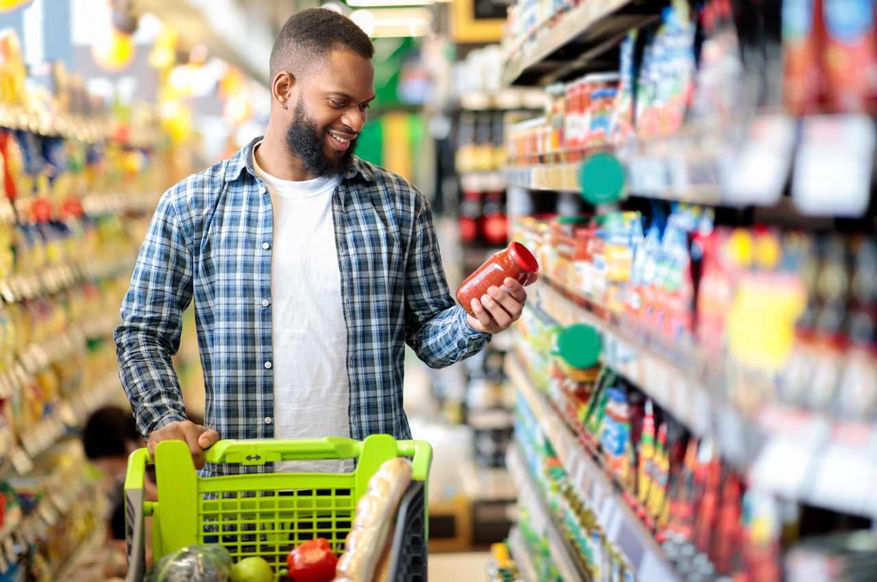 Homem sorridente realizando compras no supermercado e verificando o preço do molho de tomate na prateleira.