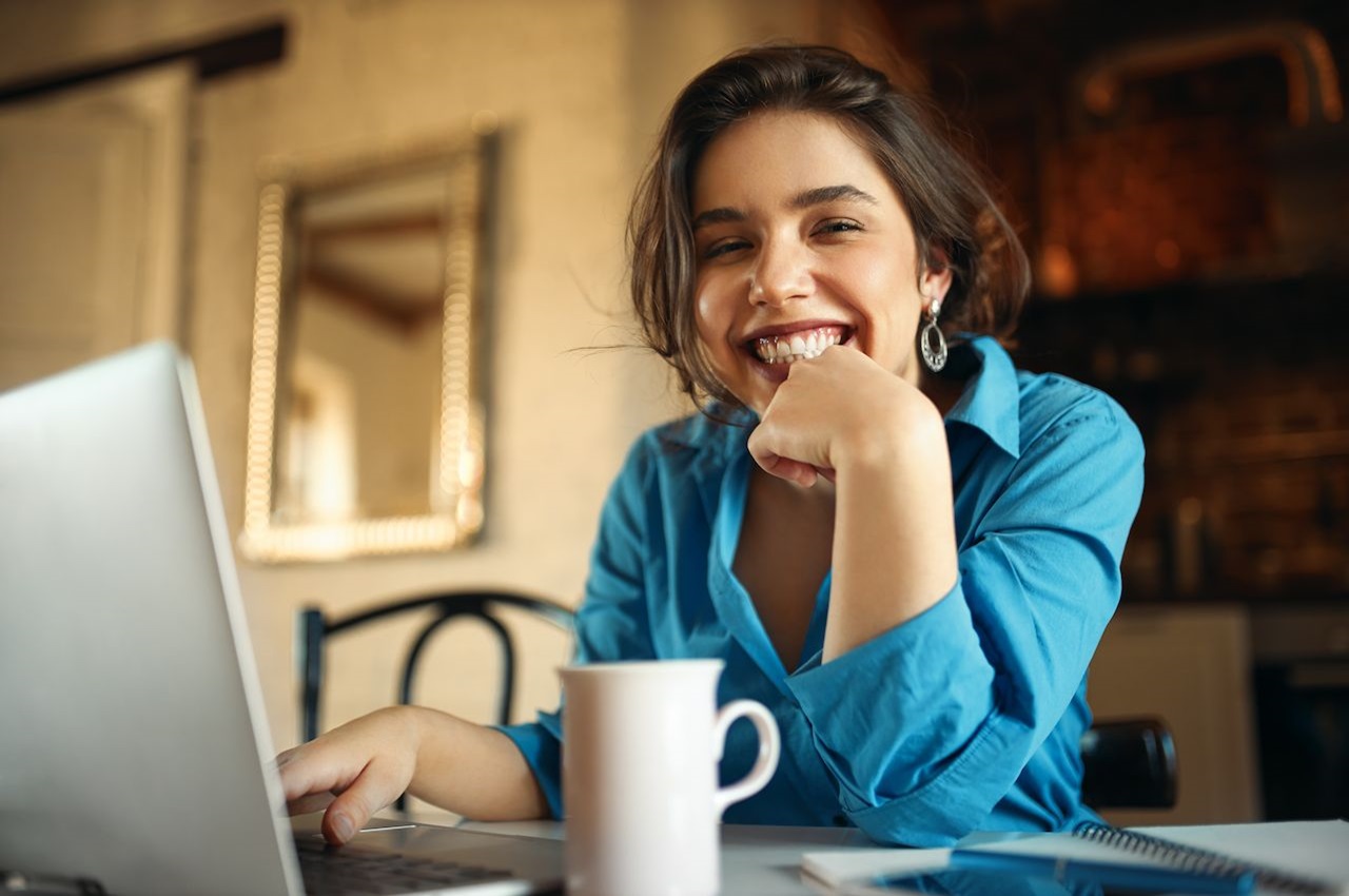 Mulher sorridente de cabelos castanhos, vestindo blusa social azul e ditando no computador da empresa.