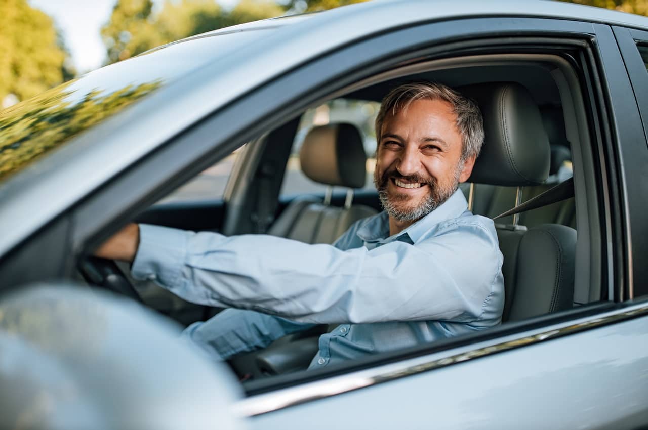 Homem sênior sorridente, dirigindo veículo de aplicativo. Ele veste blusa social azul clara.