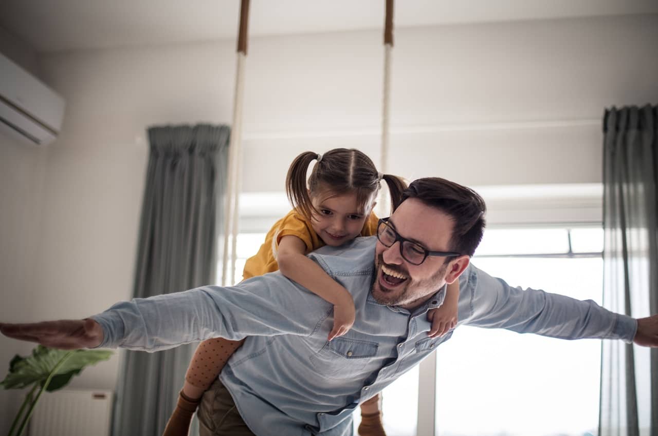 Homem e sua filha pequena com expressões sorridentes brincando na sala de estar de aviãozinho. A criança está nas costas do homem, enquanto ele abre os espaços para imitar as asas do avião.