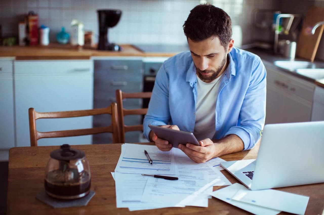 Homem com expressão concentrada segurando tablet e verificando várias contas a pagar para não ficar com nome sujo.