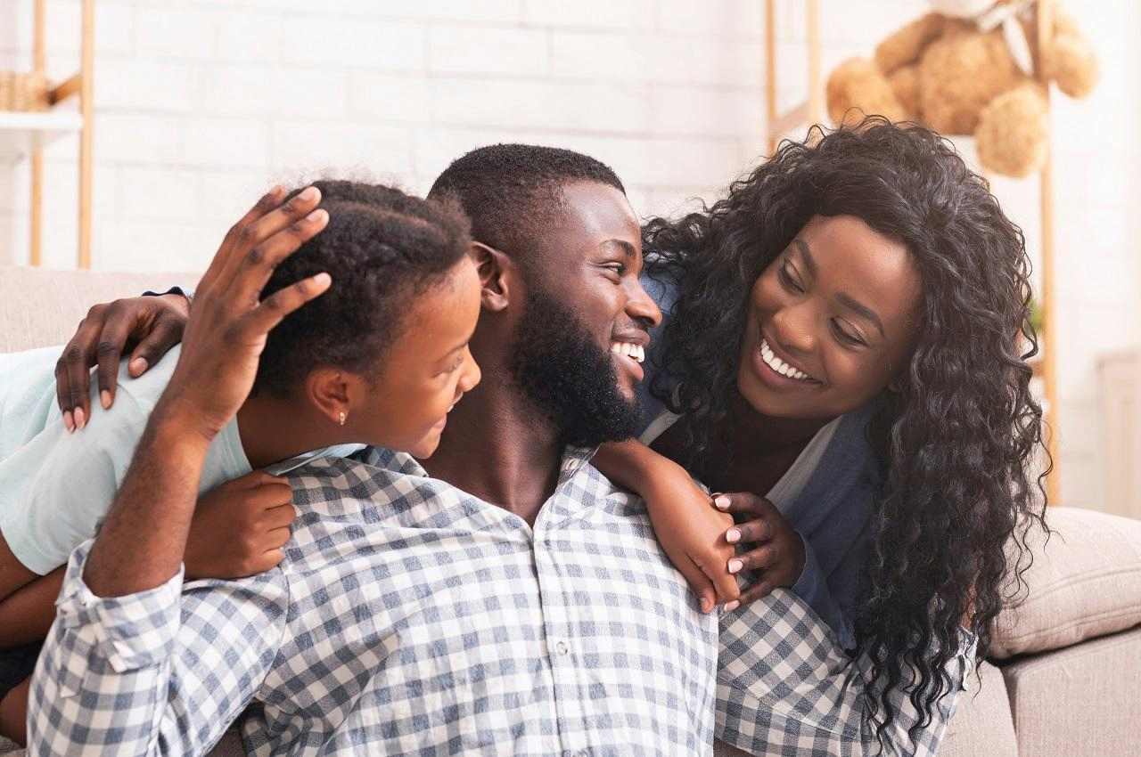 Foco em casal (homem e mulher sorridentes) e seu filho pequeno abraçados em cima do sofá. Eles sorriem um para outro.