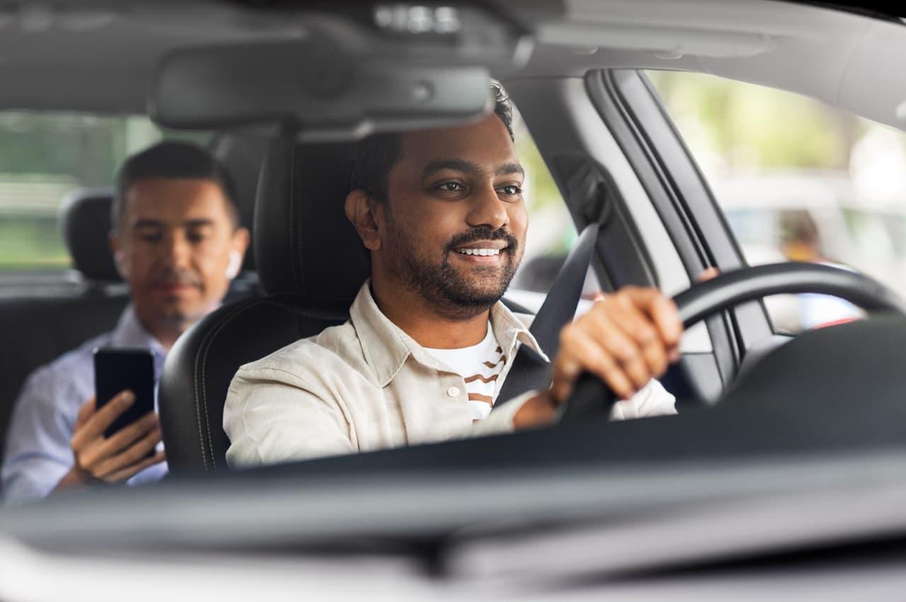 Motorista de aplicativo homem dirigindo seu veículo e transportando passageiro no banco de trás do carro. O passageiro acompanha sua viagem pelo celular.