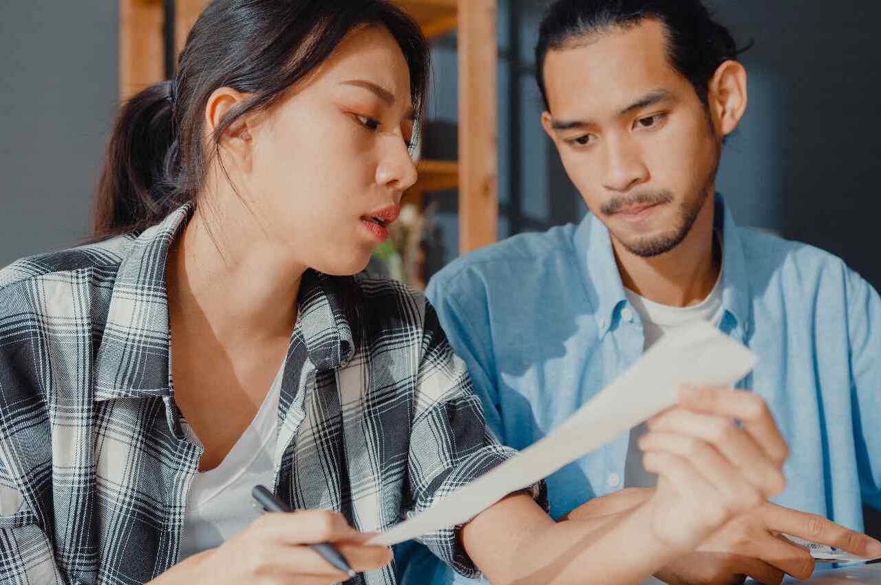 Mulher com expressão surpresa, segurando nota fiscal de gastos e mostrando para um homem ao seu lado.