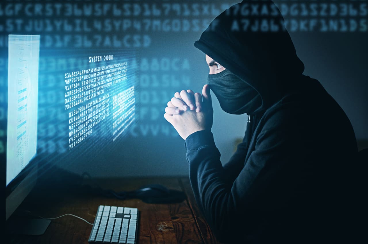Homem vestindo capuz preto e máscara enquanto olha para um monitor com códigos digitais sobrepostos em um ambiente escuro, simbolizando o golpe da falsa polícia federal.