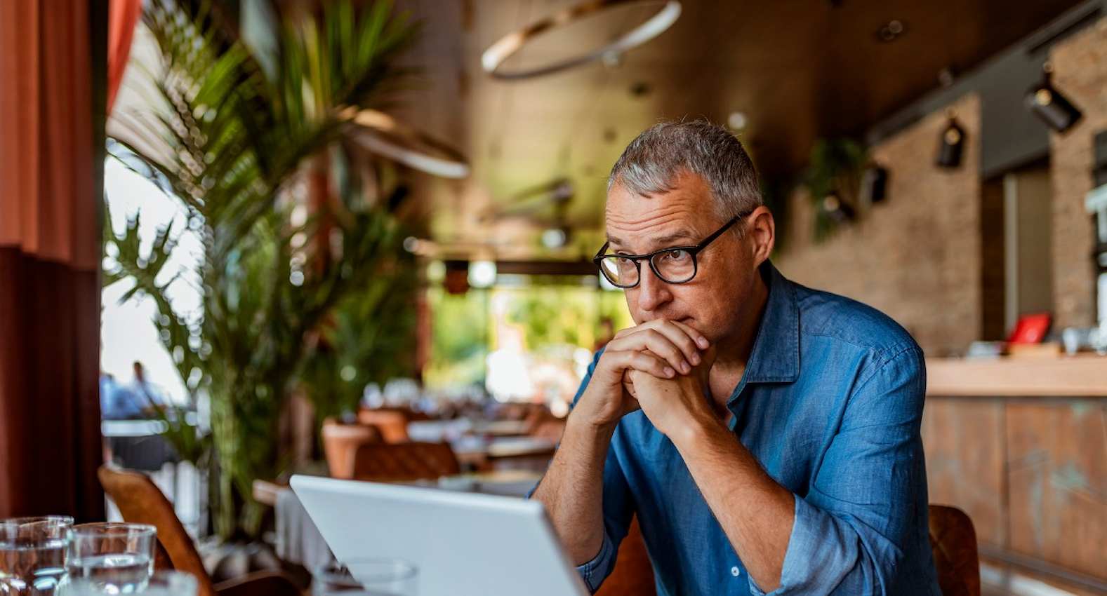 A imagem mostra um homem, sentado à uma mesa, usando seu laptop. Ele aparenta estar preocupado.