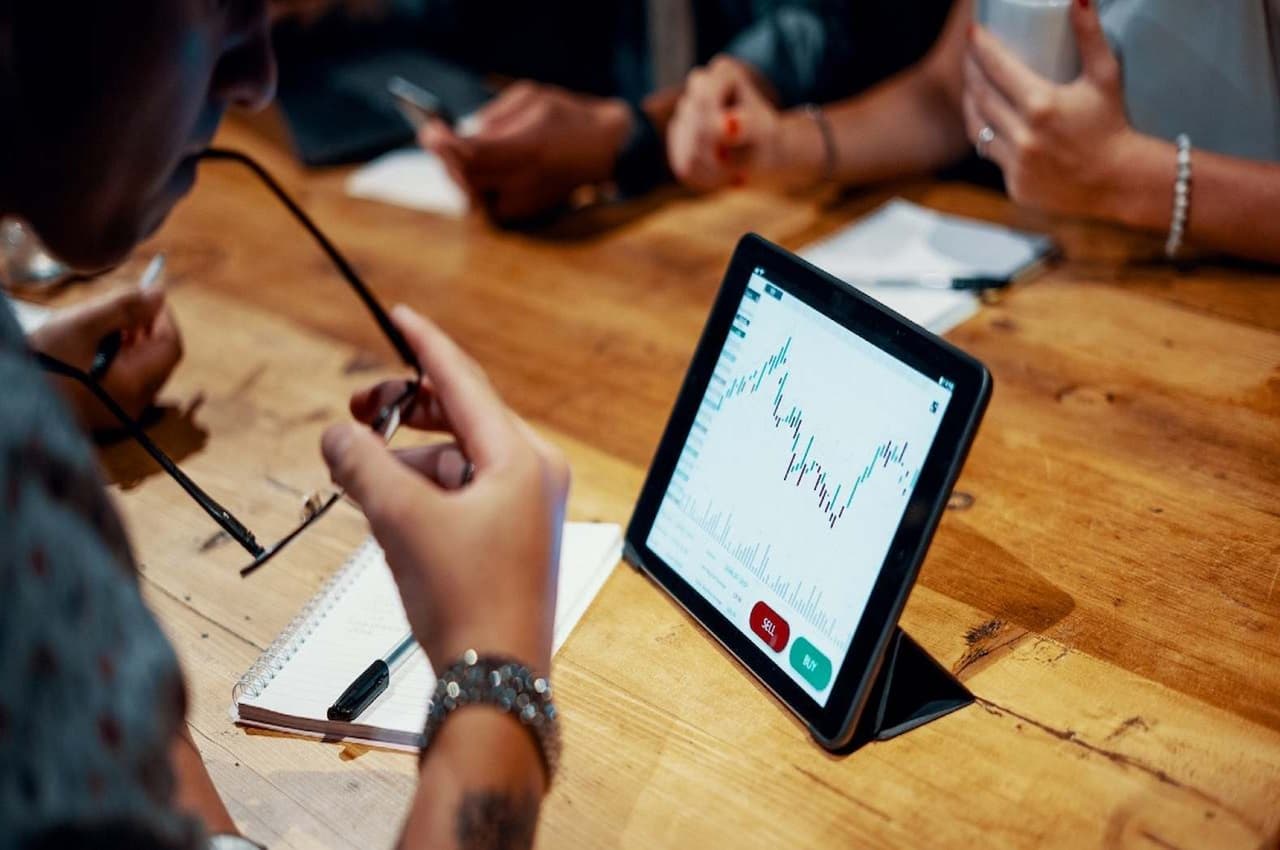 Mesa de escritório com pessoas ao redor analisando um gráfico instável de metas financeiras no tablet.