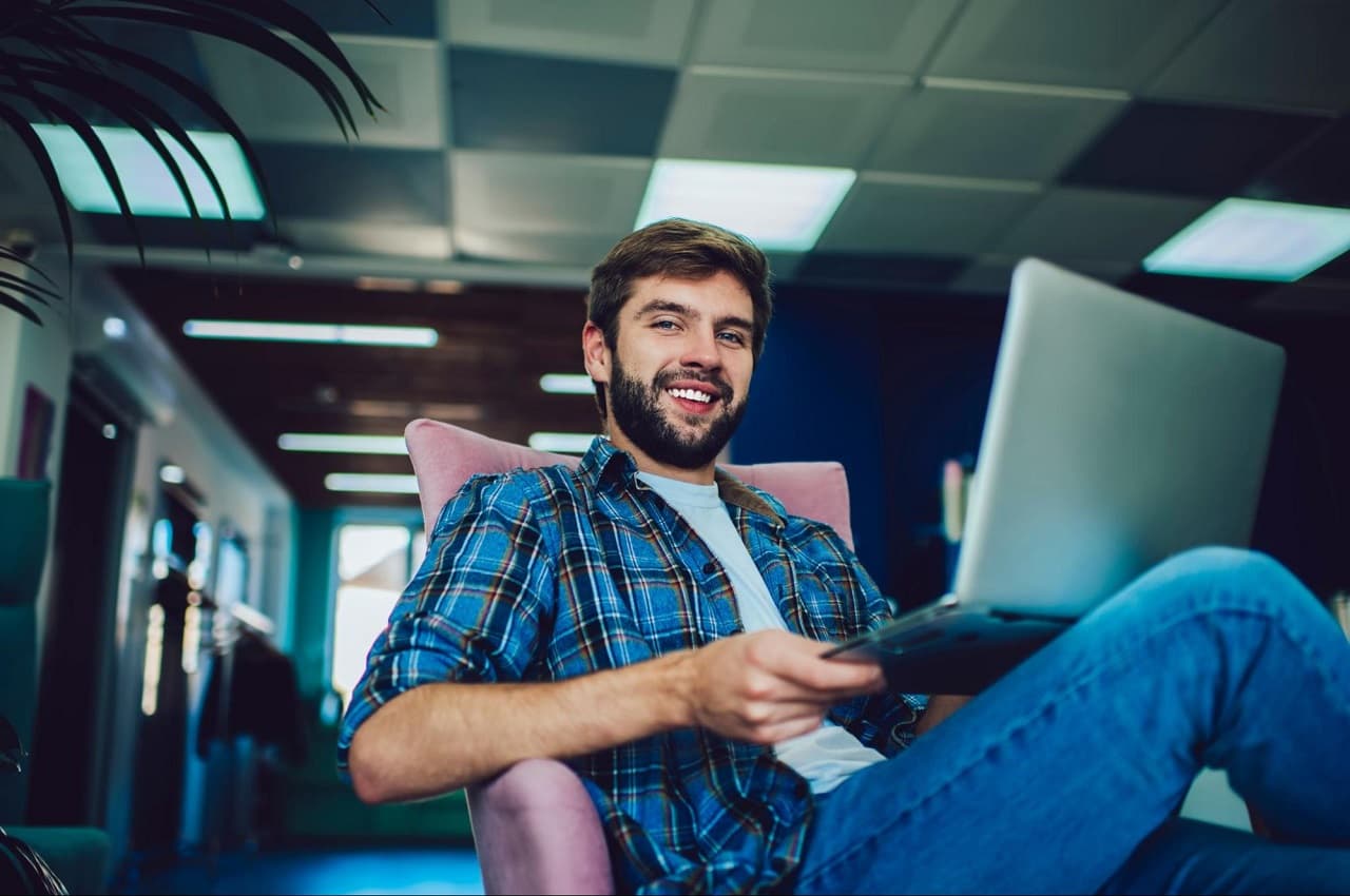 Homem sorridente, vestindo camisa xadrez azul e calça jeans, sentado na cadeira de escritório e segurando um computador no colo.