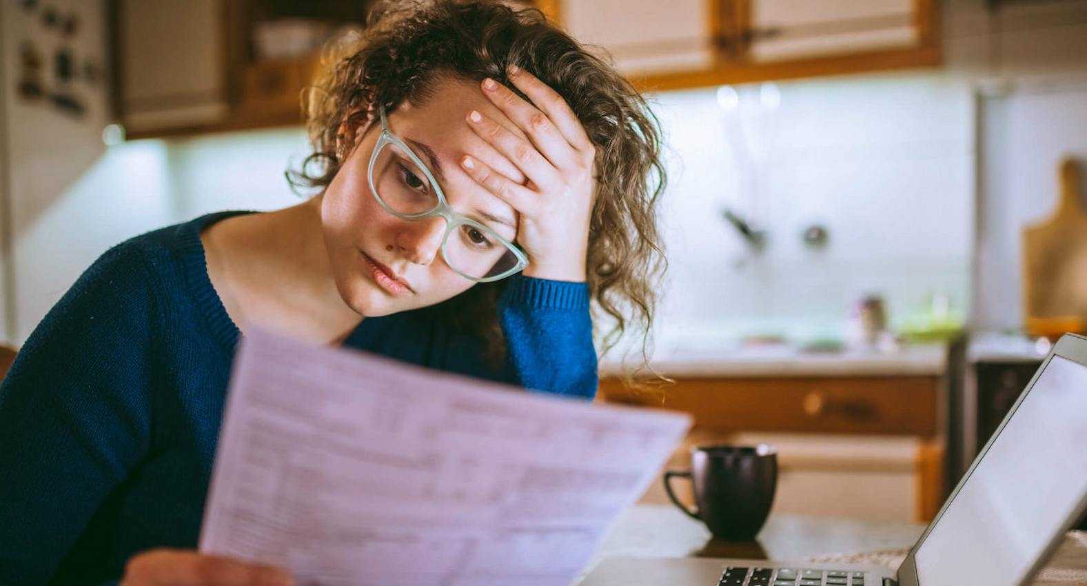 A imagem mostra uma mulher, com aparência de preocupada. analisando um boleto. À sua frente, seu laptop.