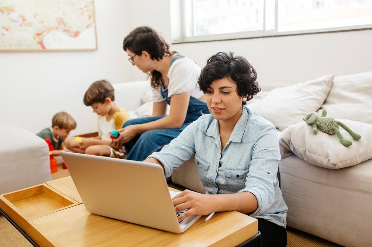 Casal de mulheres cuidando de duas crianças pequenas na sala de estar. Uma das mulheres está com a expressão concentrada utilizando o computador para fazer o controle financeiro familiar.