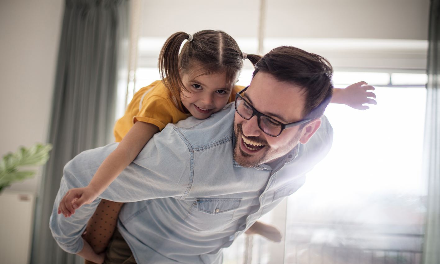 A imagem mostra um pai carregando sua filha em suas costas. Eles estão felizes.