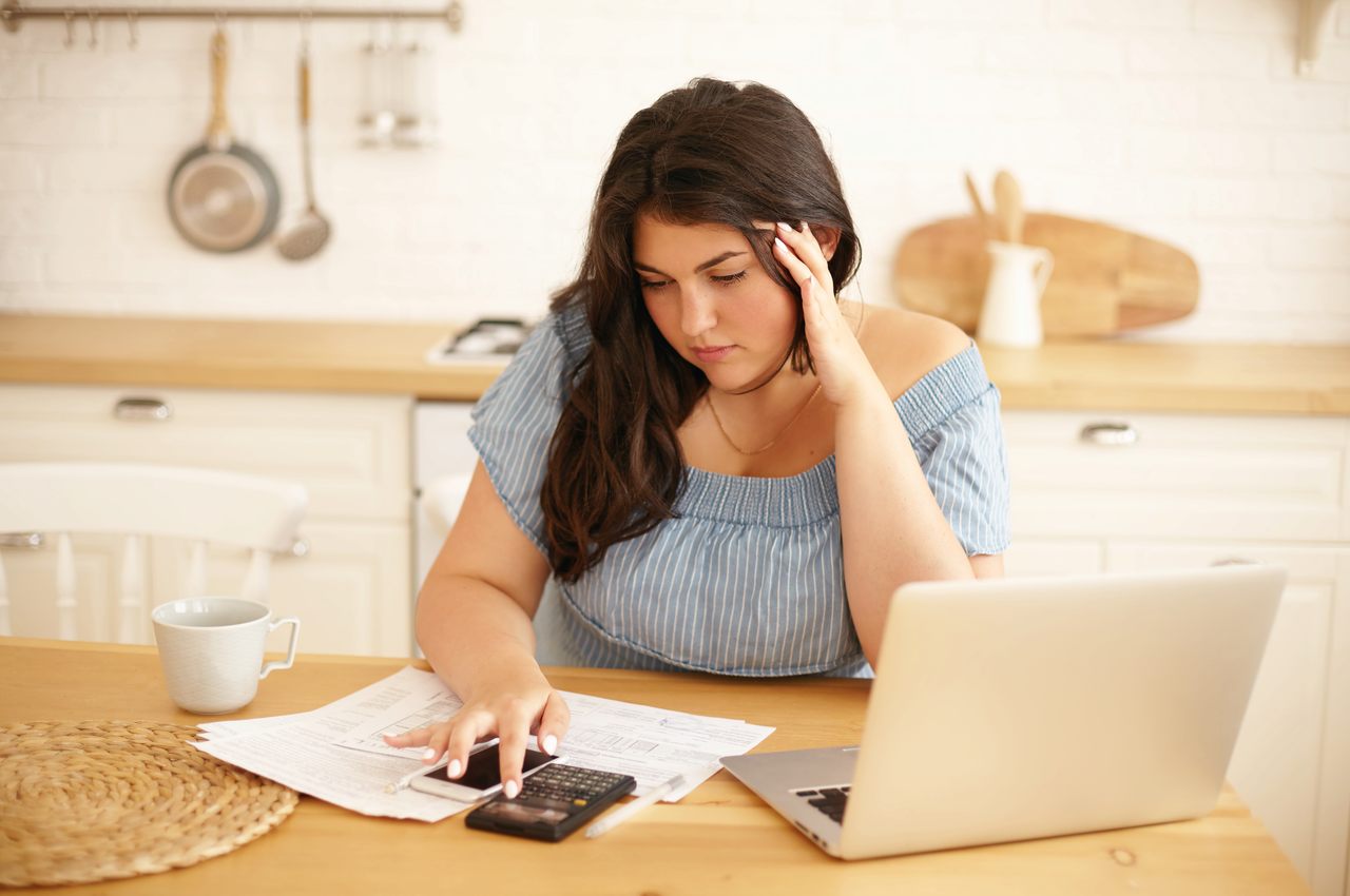 A imagem mostra uma mulher sentada a uma mesa, à frente de seu laptop. Ela está fazendo contas em uma calculadora.