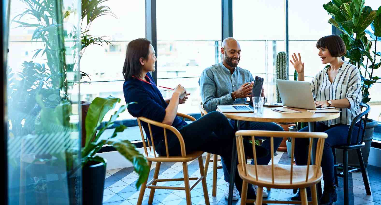 A imagem mostra três pessoas sentadas em uma mesa redonda. Duas delas estão usando laptops e conversando.