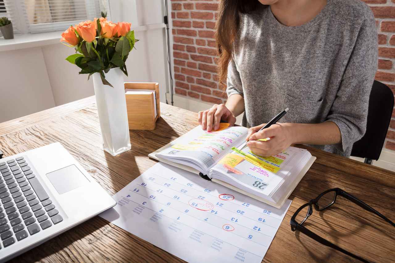 A imagem mostra uma mulher fazendo anotações em sua agenda. Ao seu lado, um vaso, um laptop, óculos de grau e um calendário.