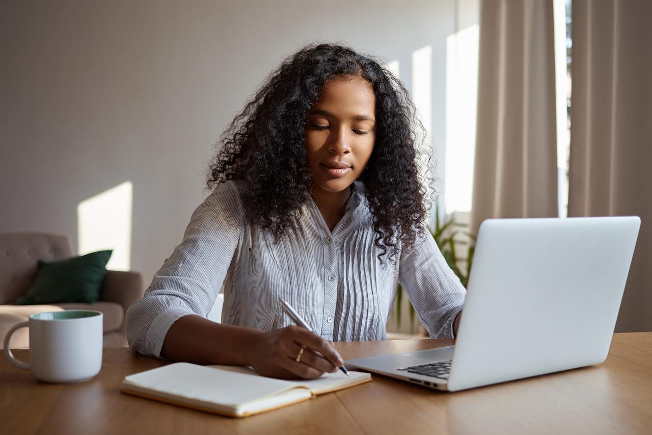 A imagem mostra uma mulher sentada à uma mesa, fazendo anotações em uma caderneta e usando seu laptop.