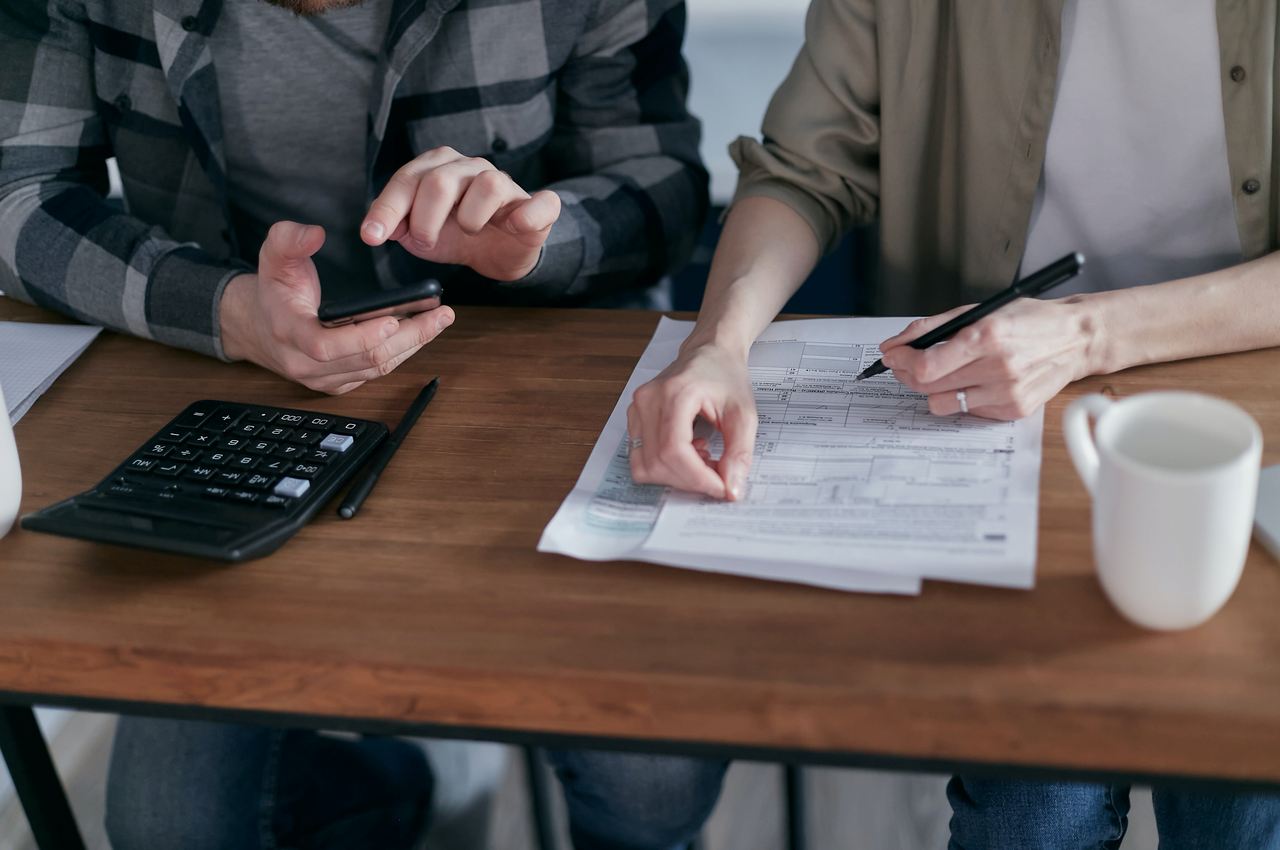 Duas pessoas conversando (seus rostos não aparecem na imagem) e olhando para contas a pagar sobre a mesa e ao lado de um calculadora comum.