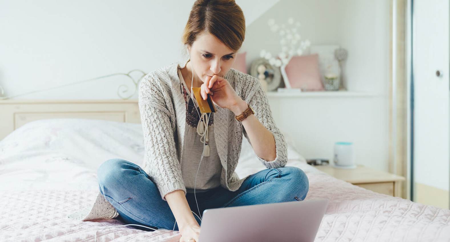 A imagem mostra uma mulher sentada a sua cama, usando seu laptop, fazendo compras em seu cartão de crédito.