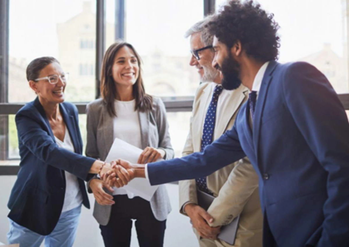 Dois homens e duas mulheres em ambiente de escritório. O homem mais jovem aperta a mão da senhora mais velha, fechando uma renegociação de dívidas.
