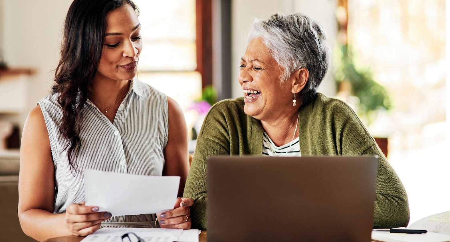 A imagem mostra duas mulheres. Uma delas está usando um laptop e a outra está com um papel na mão.