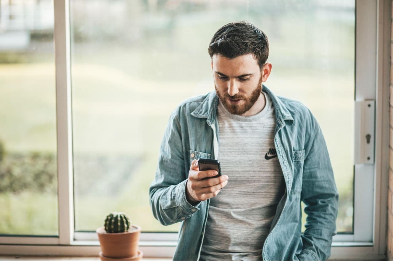 Homem com expressão concentrada apoiado no beiral de uma janela fechada e digitando no aparelho. Ele veste blusa básica cinza da marca Nike e jaqueta jeans.