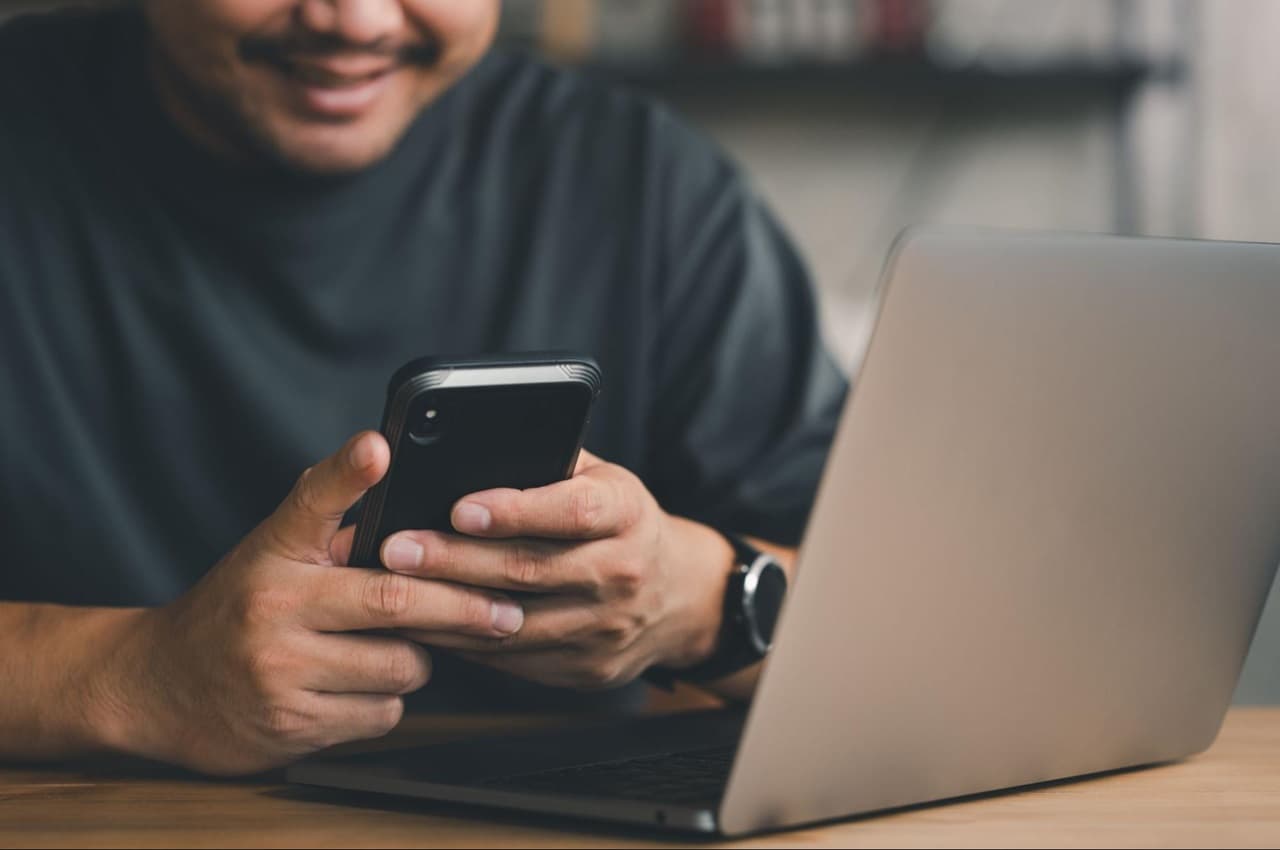 Homem sorridente segurando celular com duas mãos em cima de um computador aberto sob a mesa. A pessoa veste blusa básica preta e relógio.