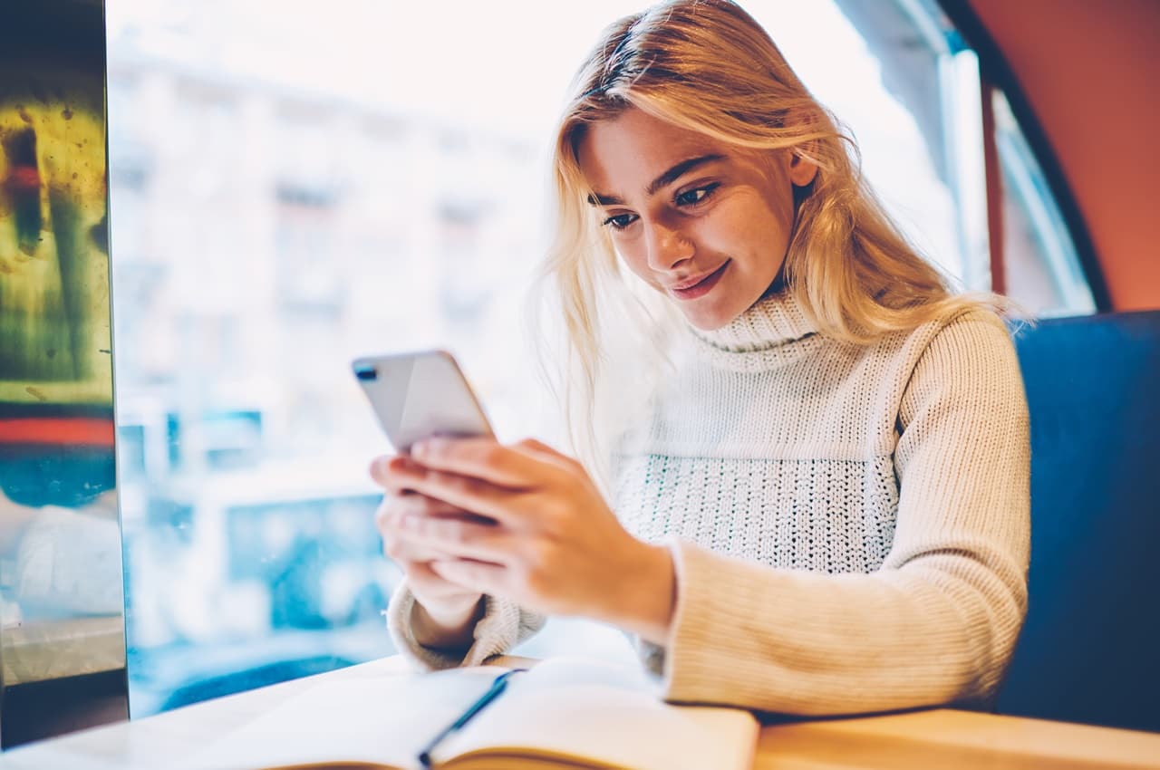 Mulher loira sorridente acessando celular para pedir empréstimo online. A mulher está sentada em uma cafeteria de banco azul e com um caderno de anotações aberto.