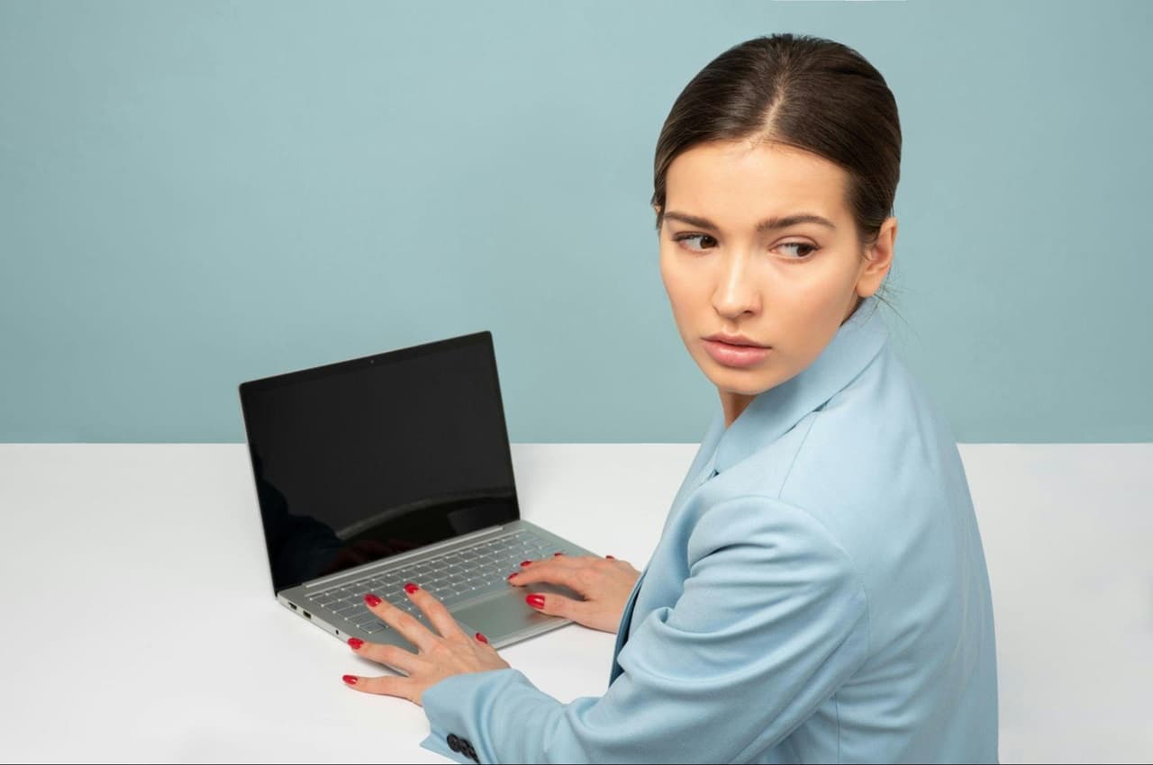 Mulher digita no laptop com a tela desligada e olha por cima do ombro com expressão concentrada. A mulher veste blusa blazer azul-claro e está sentada em frente a uma mesa branca.