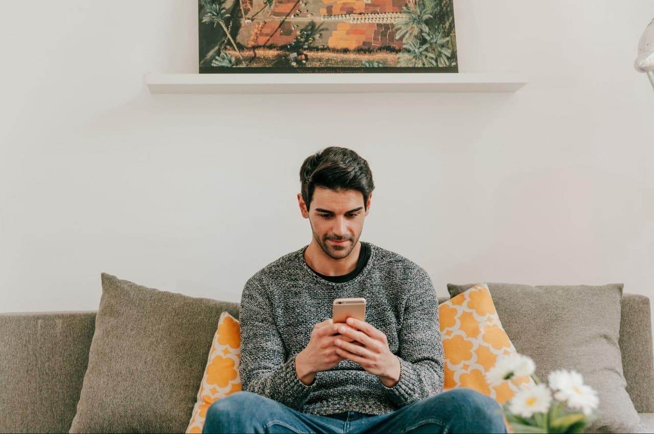 Homem sorridente sentado no sofá segurando celular nas mãos. O sofá tem duas almofadas amarelas e o homem veste calça jeans e suéter cinza.