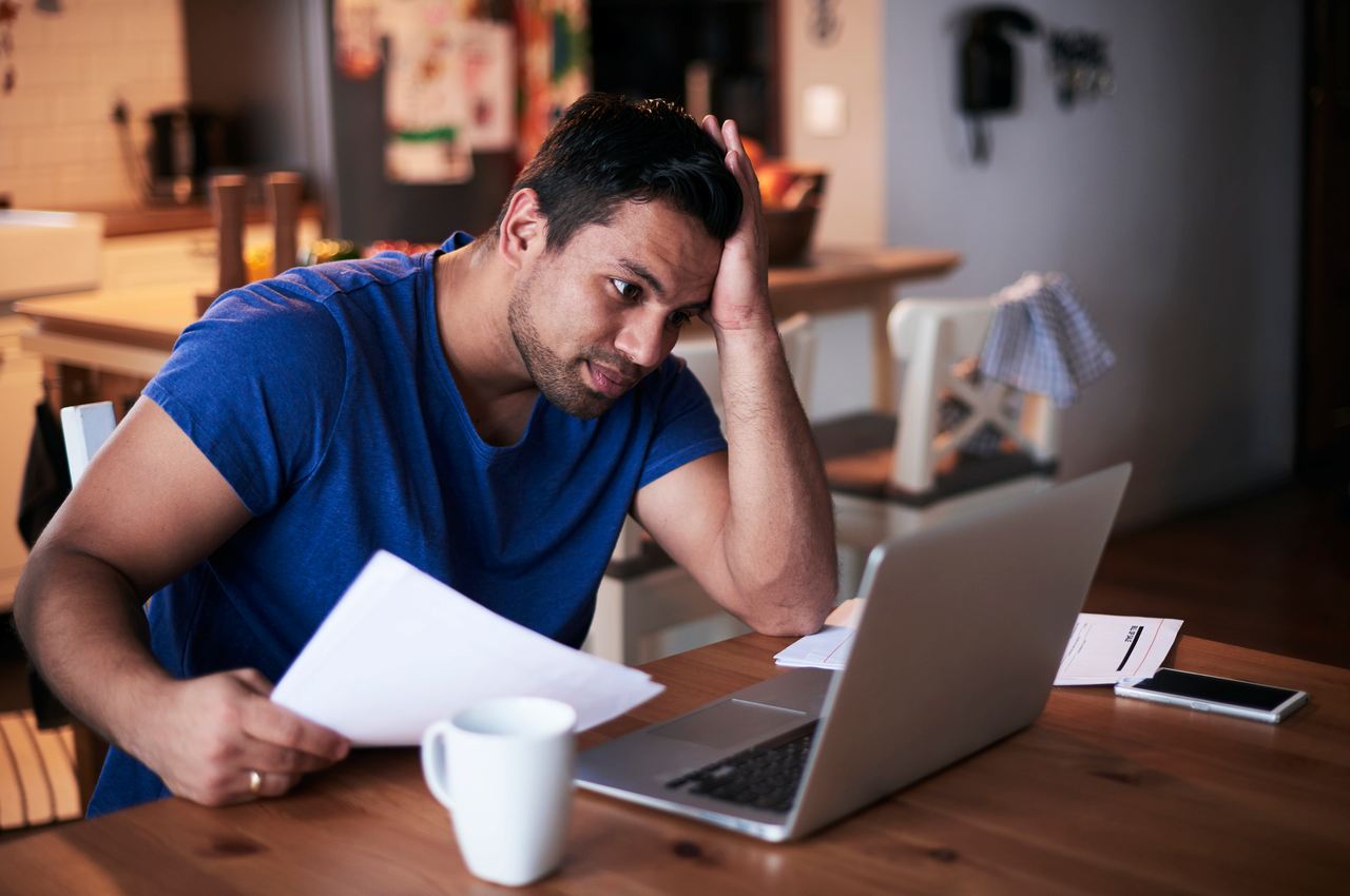 Homem com expressão concentrada apoiando a mão direita na cabeça e segurando papéis com a mão esquerda, enquanto pesquisa no computador golpes na internet.