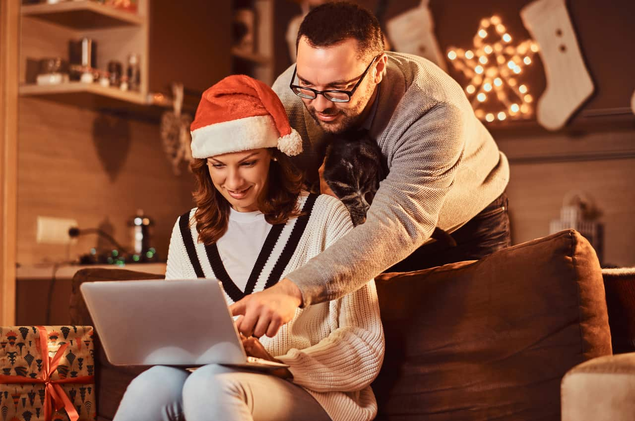 Mulher sorridente com touca de papai-noel, sentada no sofá e utilizando o computador, enquanto um homem sorridente aponta para a tela do aparelho.