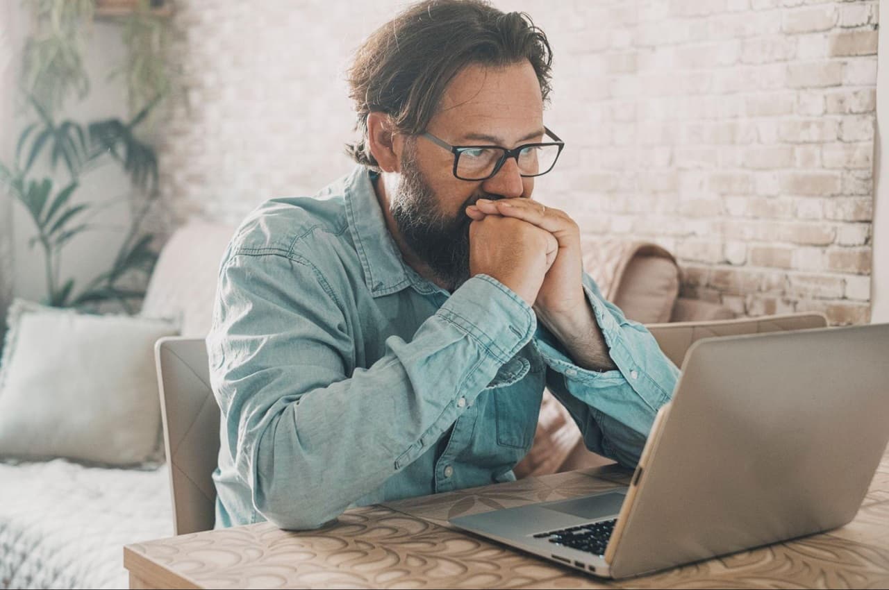 Homem com expressão concentrada, apoiando as mãos fechadas na boca e olhando para tela do computador. Ele está na sala de estar, veste blusa jeans de botões e óculos de grau.