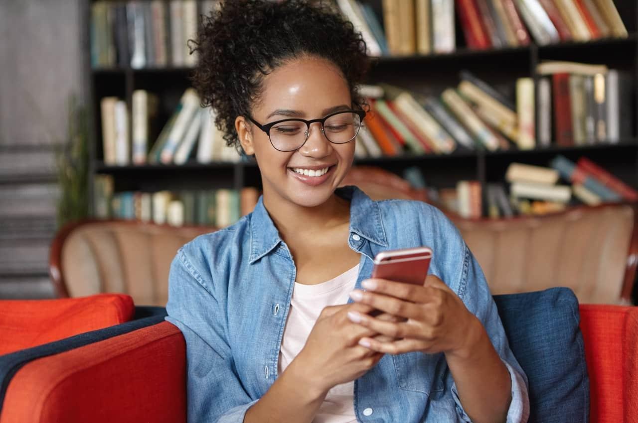 Mulher sorridente sentada em sofá vermelha, usando celular para acessar canais oficiais e não cair em golpes. A pessoa usa óculos, veste camisa branca básica e jaqueta jeans aberta.