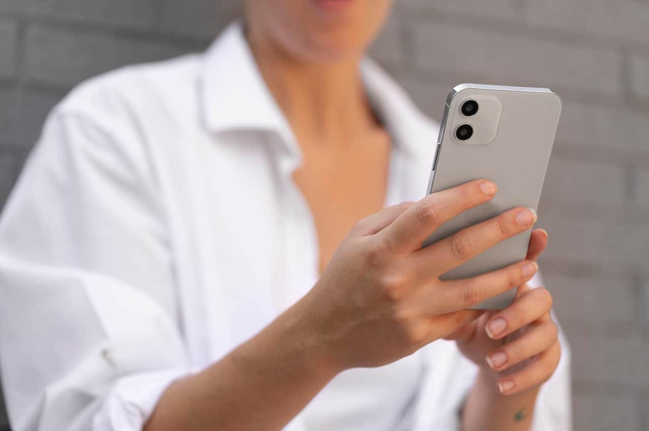 Mulher vestindo blusa social branca e segurando celular com as duas mãos na rua. O rosto da pessoa não aparece na imagem.