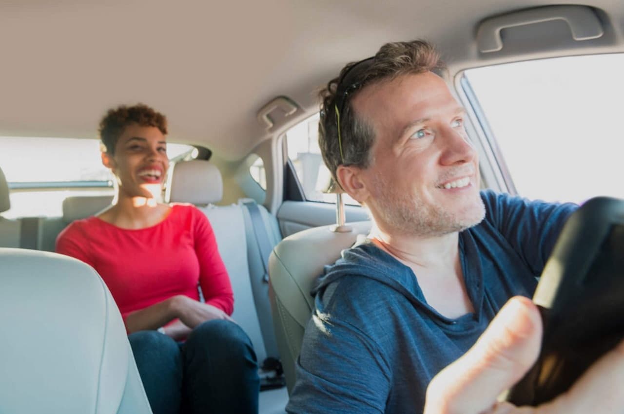 Motorista de aplicativo com expressão sorridente dirigindo um veículo e levando uma passageira mulher também sorridente e no banco de trás.