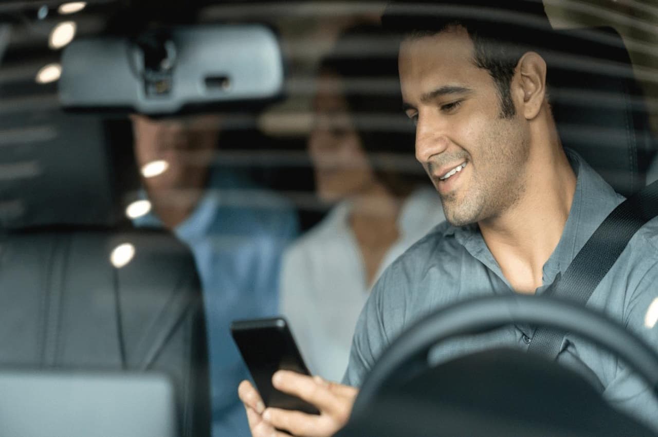 Motorista de aplicativo aceitando a viagem pelo celular e um casal de passageiros no banco de trás do veículo.