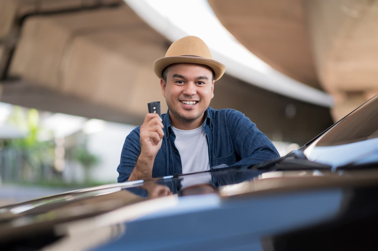 Homem sorridente, vestindo chapéu de palha e segurando a chave do carro. Ele está em pé encostado na lataria do veículo.