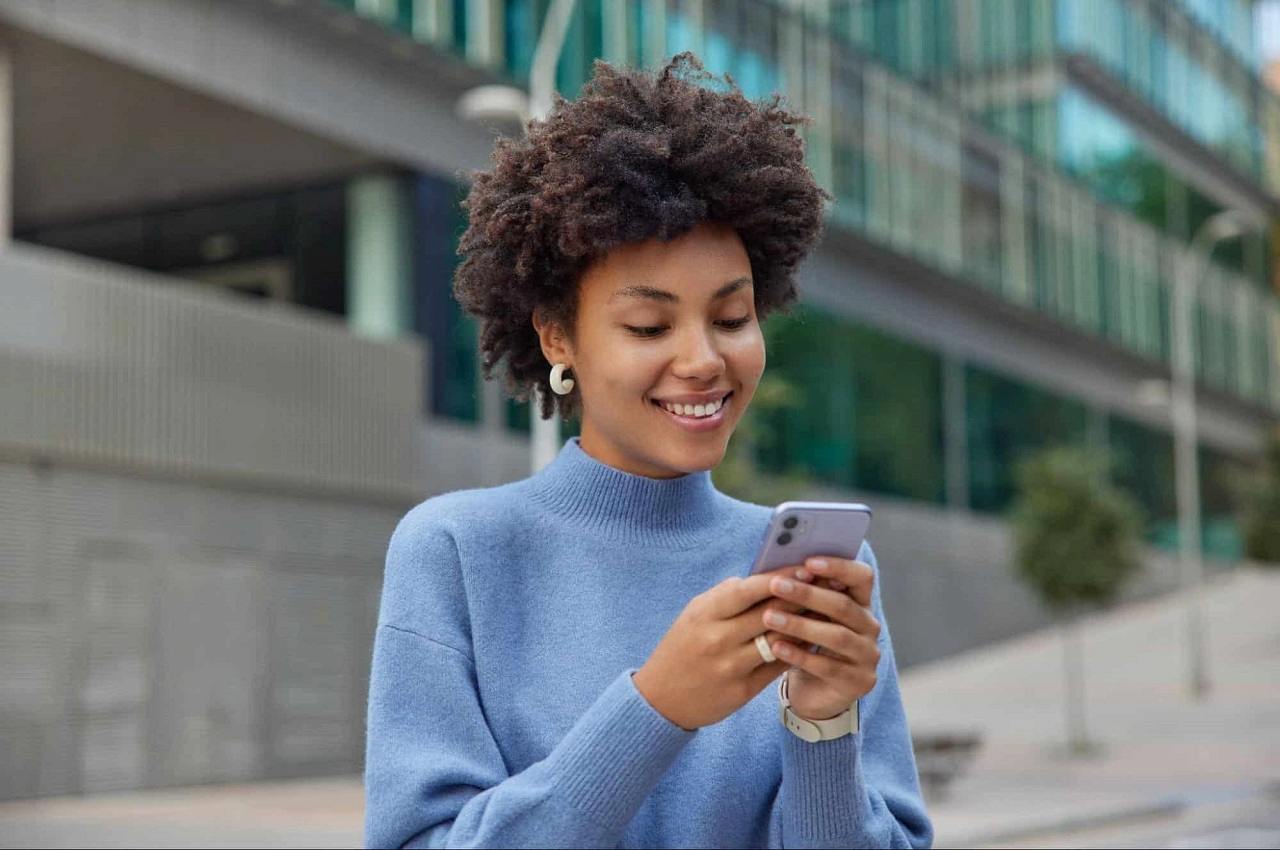 Mulher negra sorridente, vestindo blusa de gola azul e digitando no celular em frente a um prédio.