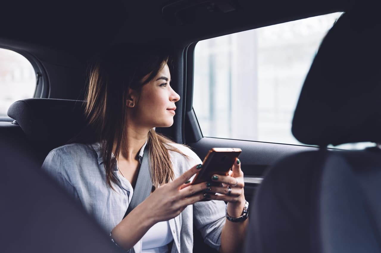 Mulher com expressão tranquila no banco passageiro do veículo, segurando um celular com as mãos e olhando a paisagem pela janela.