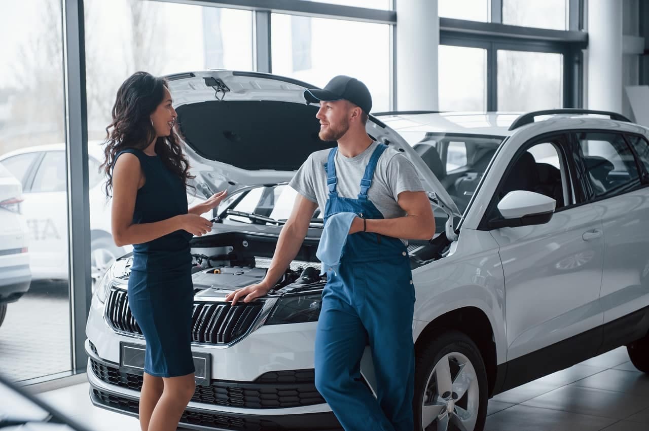 A imagem mostra uma mulher e um funcionário de uma concessionária de financiamento de carros seminovos, ao lado de um carro com capô aberto.