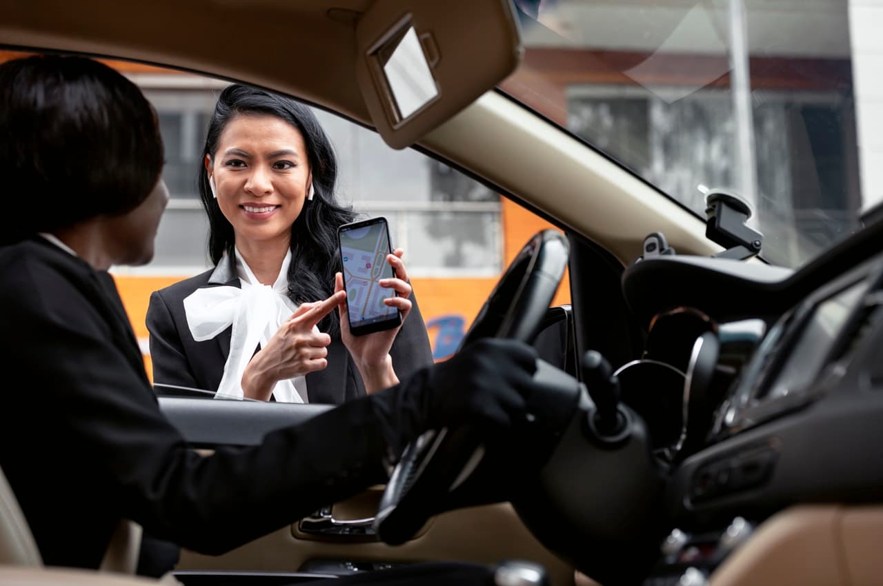 Motorista de aplicativo mulher conversando com passageira sorridente que aguarda para fora do veículo e mostra tela do celular com GPS marcando o destino da viagem.