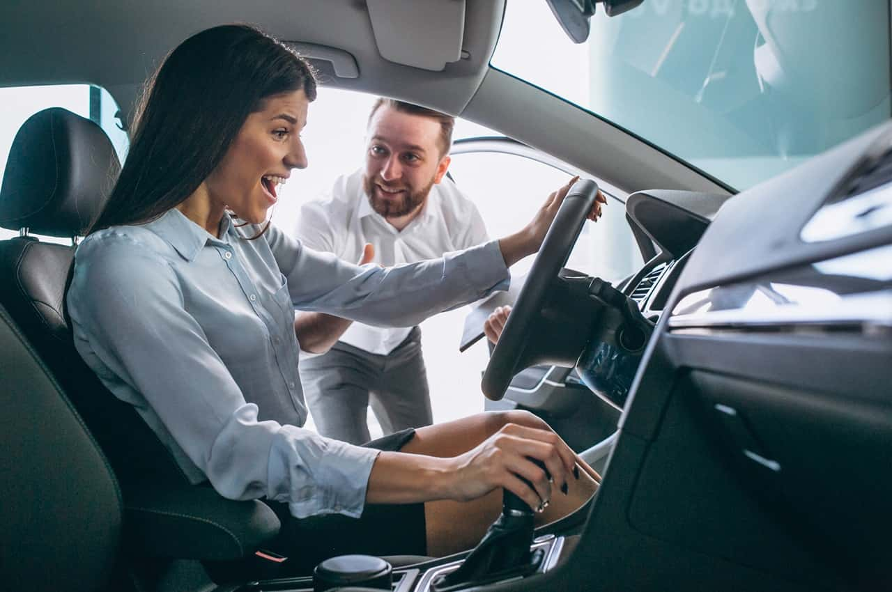 Mulher com expressão surpresa realizando teste drive-in de veículo ao lado do vendedor, para comprar carro à vista ou financiado.