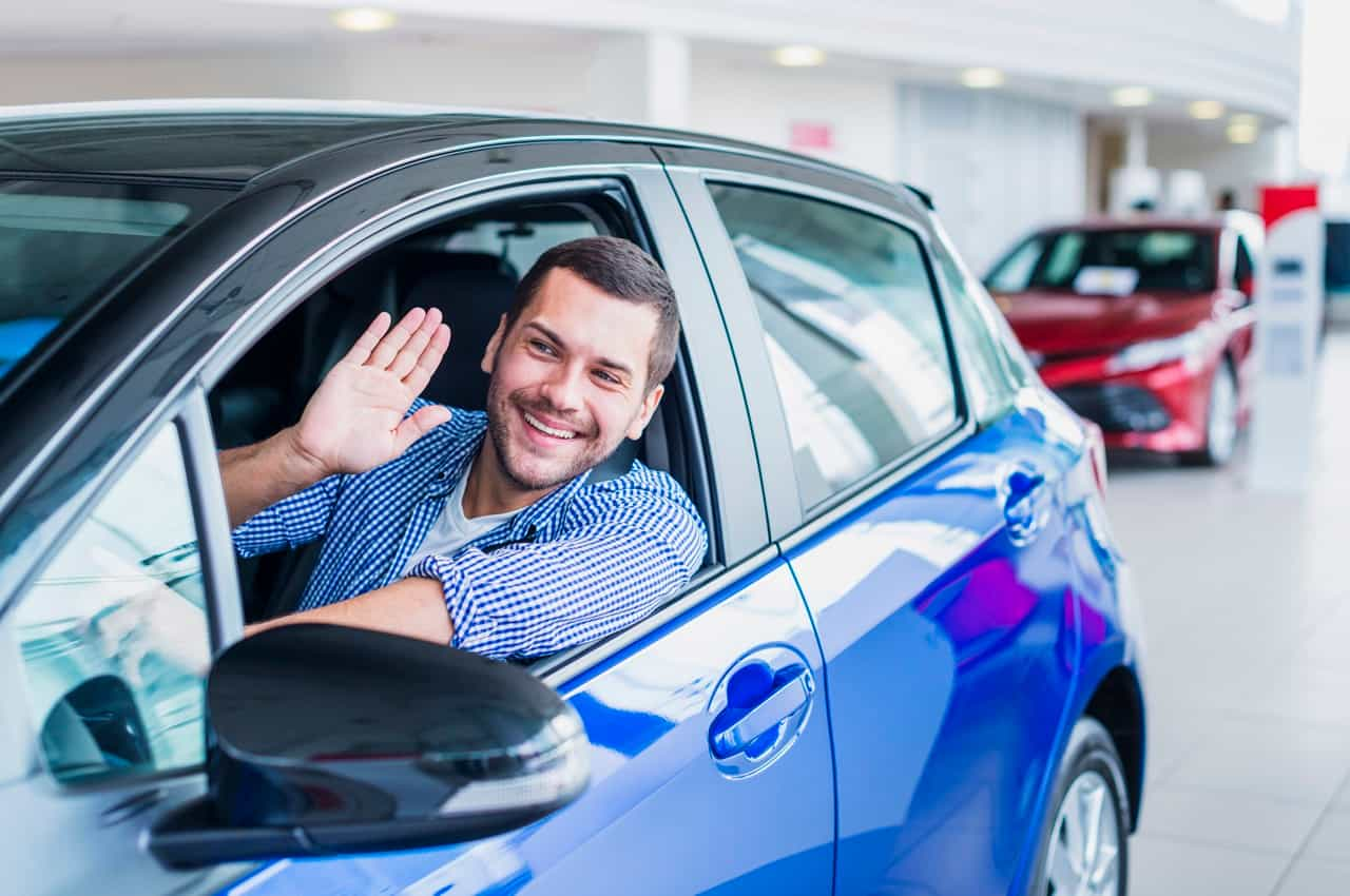 Homem sorridente dando tchau com a mão saindo de uma concessionário após comprar veículo seminovo azul.