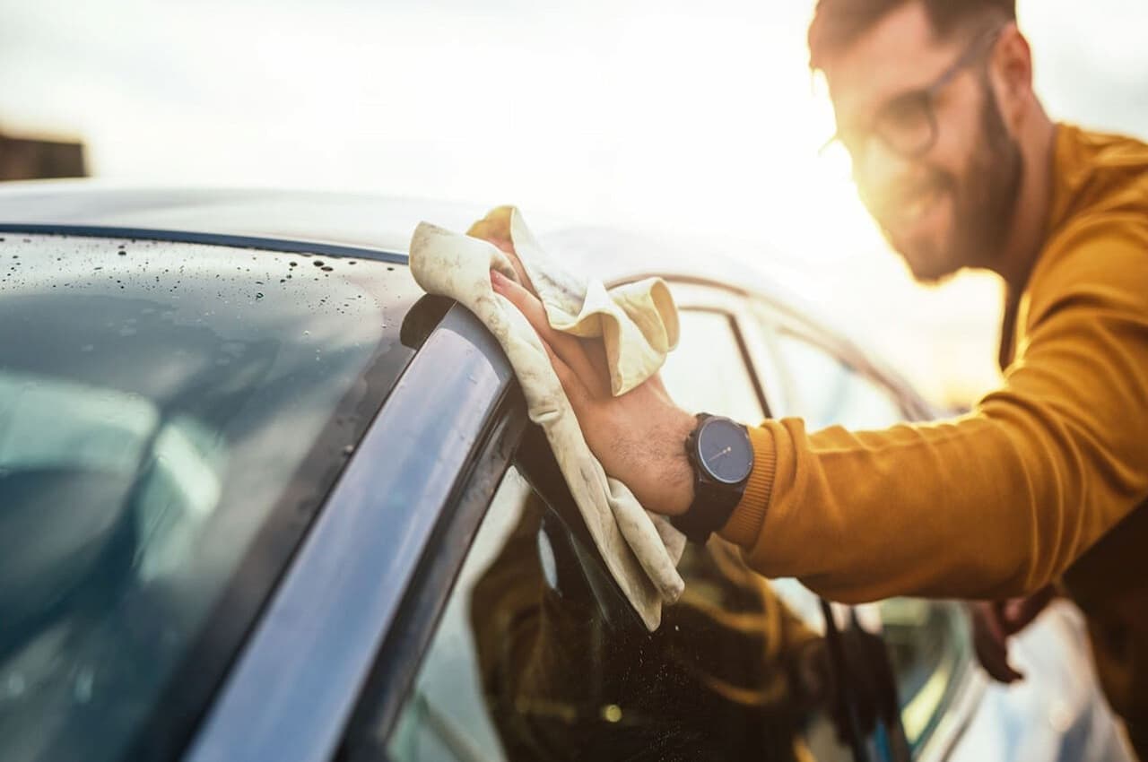 Homem sorridente secando com flanela um veículo molhado para cuidar do carro.