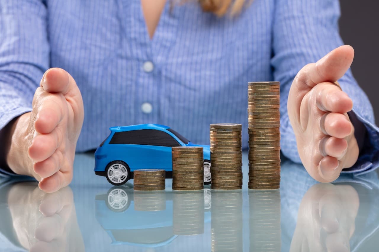 Miniatura de carro azul ao lado de pilhas de moedas e uma pessoa com as mãos envolta neles.