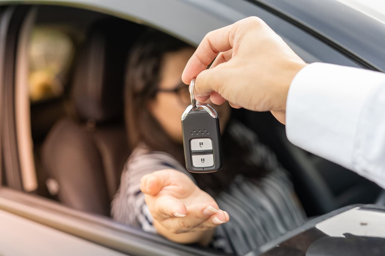 Homem (seu rosto não aparece na imagem) entregando a chave de um veículo para uma mulher que está no banco do motorista.