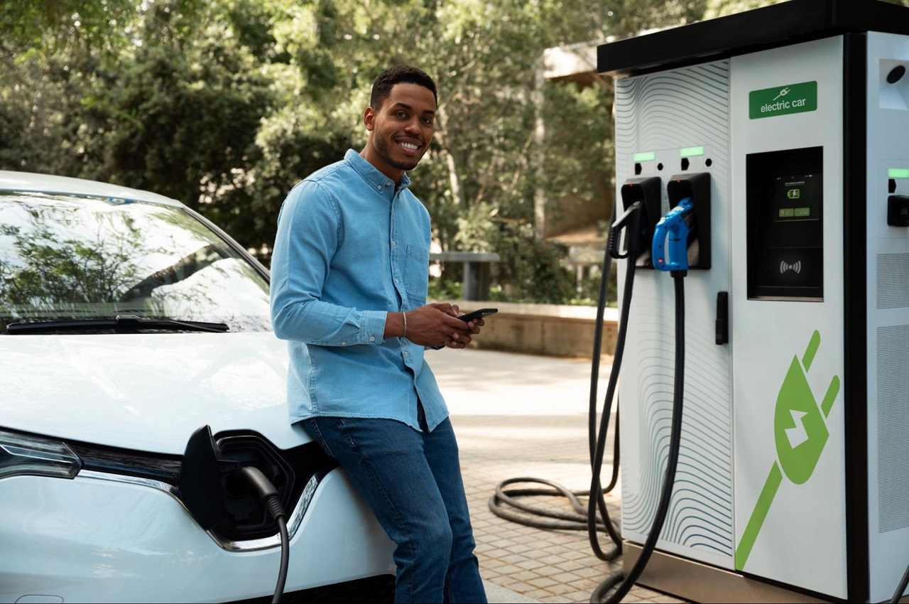 Homem sorridente com celular na mão e sentado em cima do capo do seu veículo elétrico que está estacionado reabastecendo no posto de gasolina.