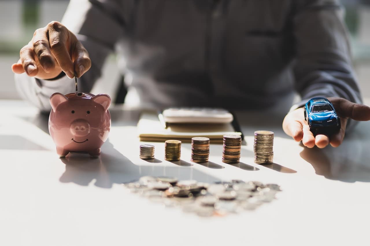 Pessoa (seu rosto não aparece na imagem) guardando moedas em um cofrinho ao lado de pilhas de moedas, economizando para comprar um carro.