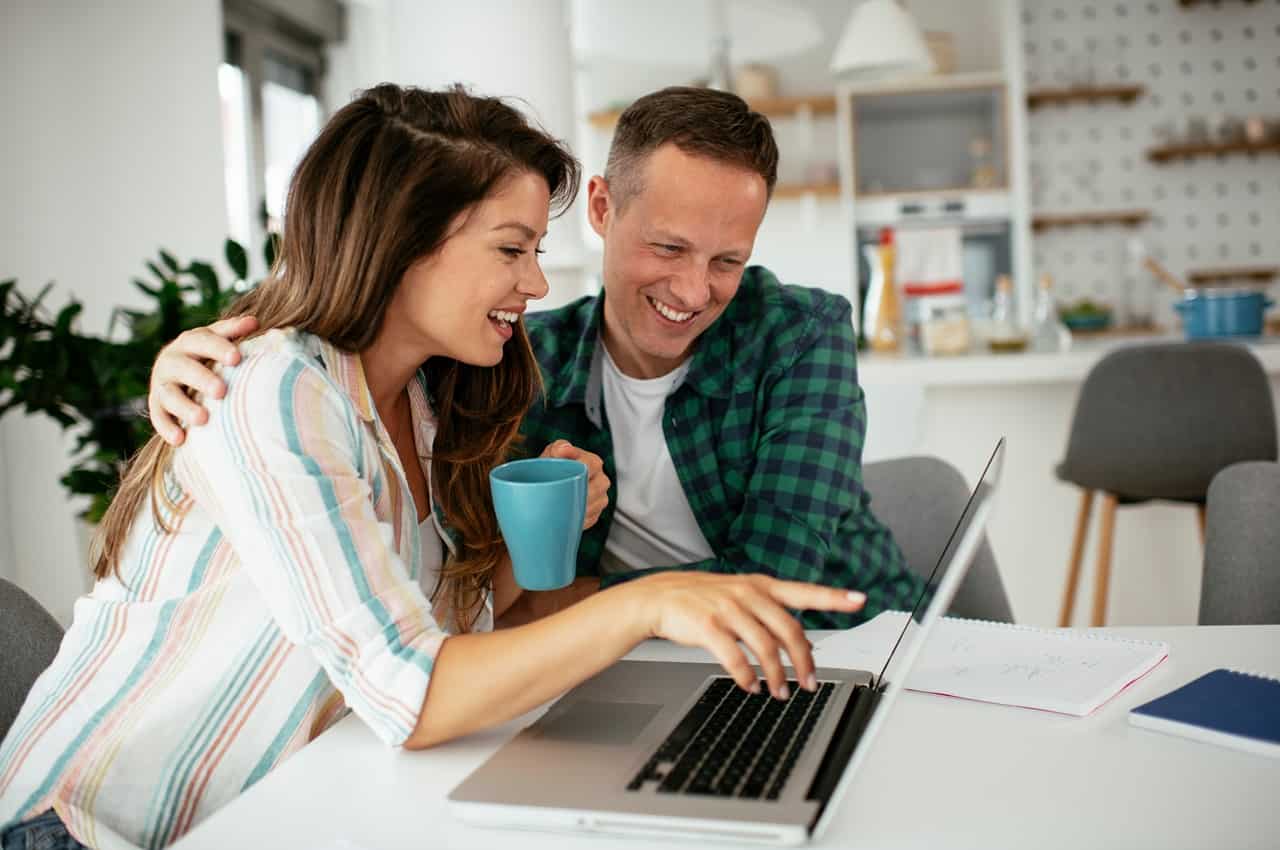 Mulher sorridente com xícara de café azul e apontando para a tela de um computador, enquanto seu companheiro sorri e olha para o aparelho.