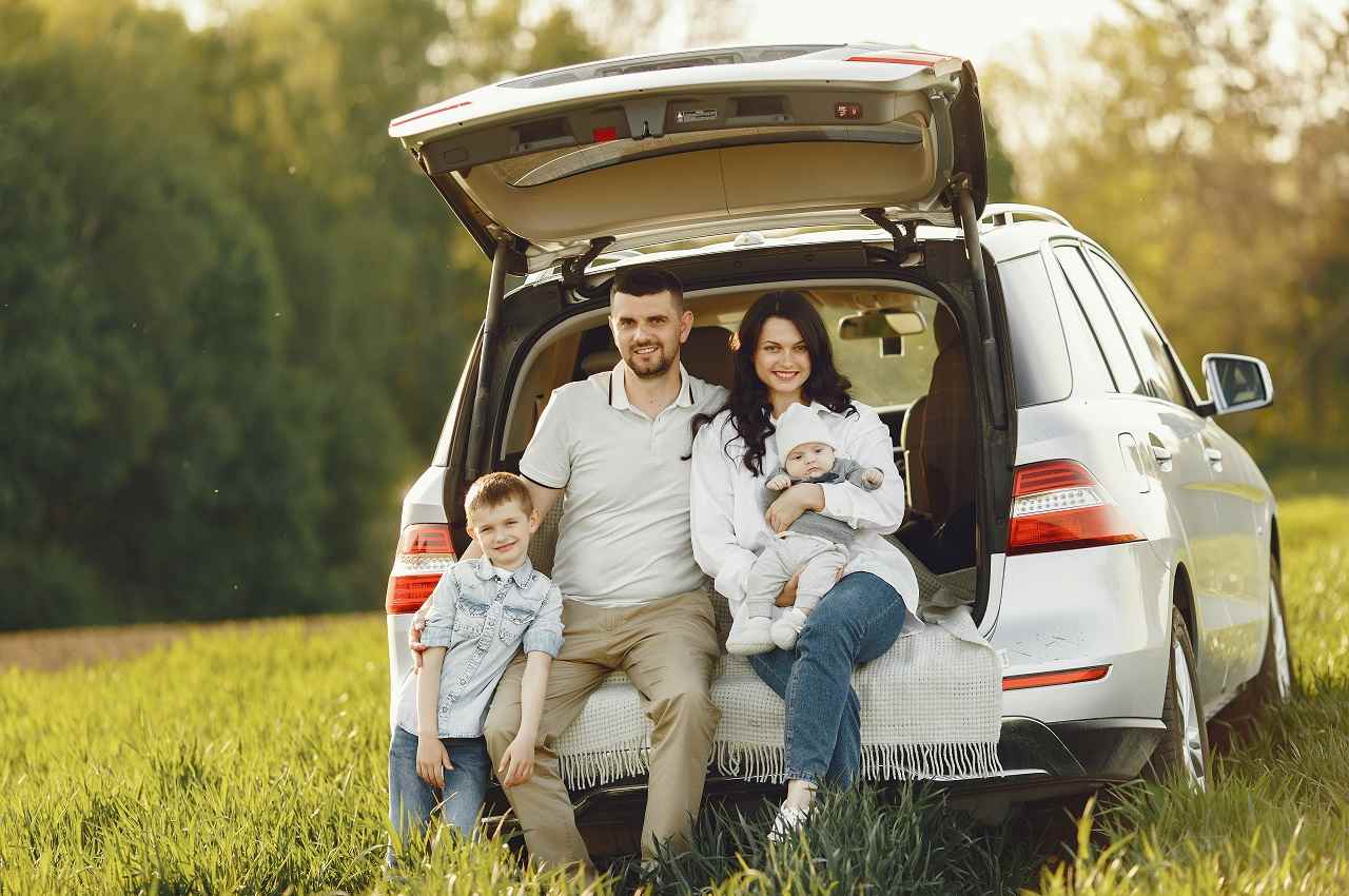 Família composta por duas crianças pequenas, mulher e homem. Eles estão sentados no porta-malas aberto do carro da família que está estacionado em um parque arborizado.